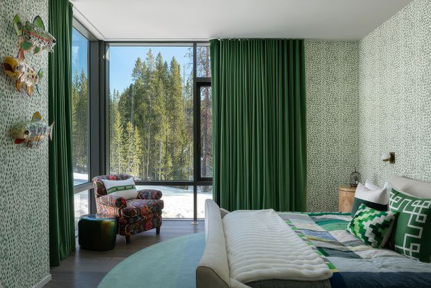 A bright, highly patterned bedroom with a large bed in the foreground, colourful geometric and patchwork bedding, and a vivid green curtain. Floor-to-ceiling corner glazing overlooks a forested landscape, while patterned wallpaper and colourful decorative wall pieces create an eclectic, playful interior. A patterned armchair and ottoman sit beside the window.