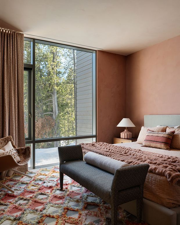 A warm, contemporary bedroom with a large floor-to-ceiling corner window overlooking trees. The room features a terracotta plaster accent wall, pale ceiling, dark window frames, a large upholstered bed with layered blush and neutral bedding, a patterned multicolour rug, a dark upholstered bench, and a small bedside table with a lamp. Textured curtains and an occasional chair add softness and visual interest.