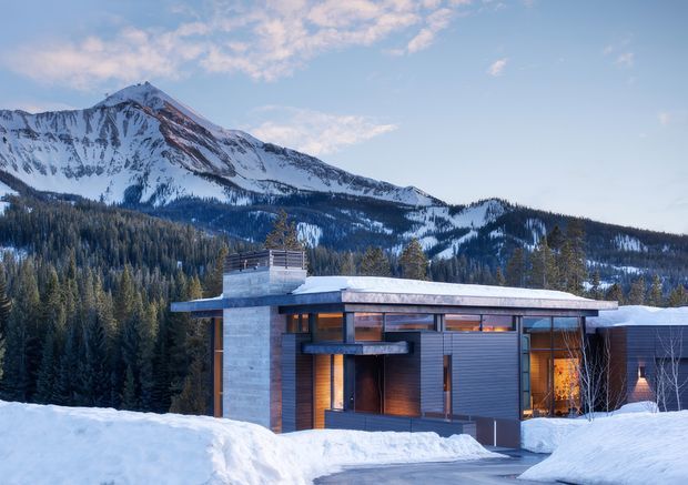 A modern flat-roofed house set in a snowy mountain landscape. The low, horizontal facade combines dark timber or wood-toned cladding, grey vertical paneling, pale stone-clad walls, extensive glazing, and a broad projecting roof. Snow covers the roof, driveway, and surrounding ground, with dense conifer forest and a prominent snow-covered mountain behind.