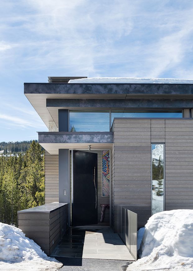 Exterior photograph of a contemporary mountain home entrance in a snowy forest setting. The facade combines dark weathered metal or rendered panels with horizontal timber cladding, broad roof overhangs, large glazed openings, and a deep black entry door. A paved walkway approaches the recessed entrance between low walls, with snowbanks and conifer-covered hills visible around the house.