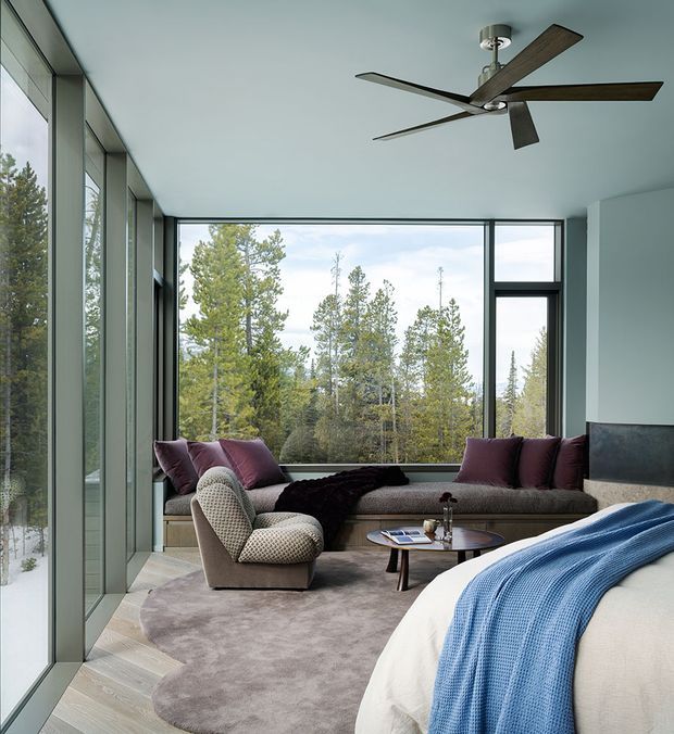 A refined open-plan bedroom-living space with floor-to-ceiling glazing overlooking a wooded landscape. The room features a large built-in window seat with deep mauve cushions, a textured lounge chair, round wood coffee table, soft grey area rug, and a bed in the foreground covered with a blue knitted throw. A contemporary multi-blade ceiling fan and dark-framed windows complement the restrained, nature-connected interior.