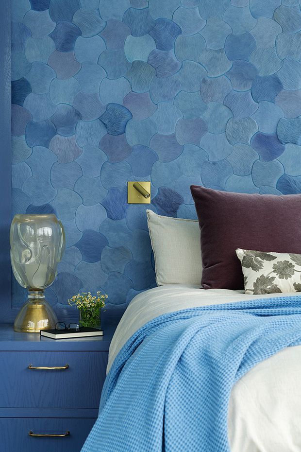 A close-up of a richly styled bedroom featuring a bed with layered white and blue bedding, a deep burgundy cushion, and a patterned accent pillow. The wall is covered in an organic overlapping scallop-shaped blue textured finish. A blue built-in bedside table with brass-toned handles supports a sculptural glass-and-metal lamp, books, and a small plant. A brass wall switch adds a contrasting detail.