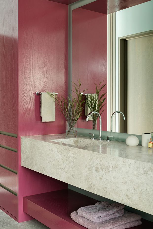 A richly coloured contemporary bathroom vanity featuring a deep burgundy-red timber-look wall and integrated open shelving, a large frameless mirror, pale stone vanity benchtop with a thick waterfall-style edge, and twin brushed metal faucets over an undermount basin. A folded towel, small vase of greenery, and neutral accessories add texture and contrast. A doorway is visible in the mirror reflection.