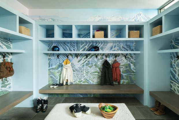 A compact, highly customised mudroom or entry storage space with pale blue built-in cabinetry, open cubbies, benches, and shelving. A patterned blue-green mural or wallpaper lines the rear wall, with woven storage baskets, hooks, coats, boots, and shoes arranged throughout. A mountain landscape band runs along the upper wall, while a central island or bench with a pale upholstered top occupies the foreground. The space is bright, playful, and strongly coordinated in cool blue and green tones.