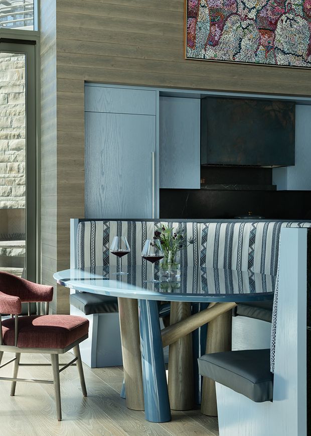A refined dining area with a built-in upholstered banquette, sculptural dining table, and coordinating dining chairs. The room features pale timber wall panelling and flooring, blue-grey built-in cabinetry, a dark fireplace surround, and a colourful framed artwork. Natural light enters through tall glazing, creating a sophisticated contemporary interior with layered textures and muted blue, grey, and burgundy accents.