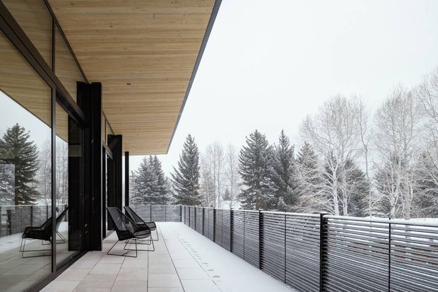 A modern covered outdoor terrace or balcony extending along a glazed building facade. The terrace has pale large-format tiled paving, a broad timber-lined soffit, black structural columns and frames, minimalist black lounge chairs, and a horizontal metal privacy balustrade. Snow-covered coniferous and deciduous trees are visible beyond the railing, creating a crisp winter setting.