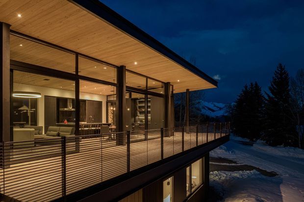 Twilight exterior view of a contemporary multi-level mountain home, focused on a broad timber-lined roof overhang and illuminated glazed living spaces opening onto a large elevated terrace with slim horizontal metal railings. The interior shows a kitchen and living area through the floor-to-ceiling glazing, while snow-covered terrain, evergreen trees, and mountain peaks form the background.