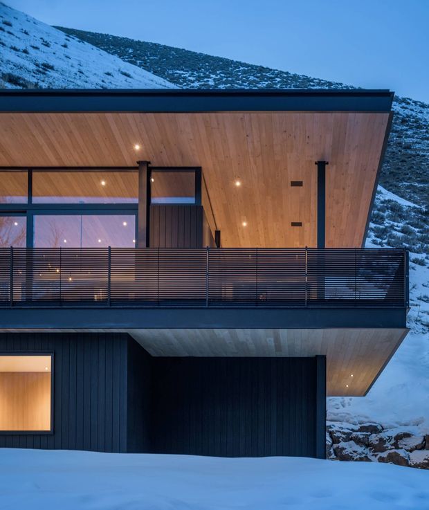 A modern multi-level mountain house viewed from outside in a snowy alpine setting. The building has dark vertical cladding, expansive glazing, a projecting upper floor, black metal balcony balustrades, and warm timber soffits and ceilings illuminated by recessed lights. Snow-covered rocky slopes surround the structure, reinforcing its rugged alpine context.