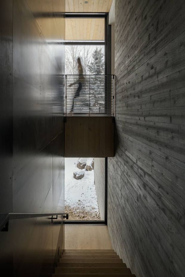 A dramatic, narrow interior stairway descending beside a textured exposed concrete wall. The stairwell has dark timber wall and ceiling panels, a timber-clad bridge or landing with horizontal metal balustrades, and tall vertical glazing that brings in views of snowy ground and trees. The space is minimalist, cool-toned, and strongly defined by its raw concrete, timber, steel, and shadowed architectural composition.