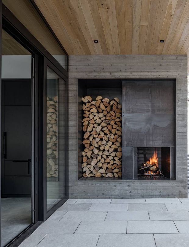 A refined modern entry or covered outdoor living area featuring a dramatic built-in fireplace and integrated firewood storage. The fireplace surround is clad in grey concrete-like panels, with a large black metal firewood recess and a smaller open firebox. A warm timber-lined ceiling contrasts with the dark-framed full-height glazed door and pale large-format tiled floor.