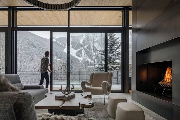 A sophisticated mountain-view living room with full-height black-framed glazing opening toward a balcony and snowy forested slopes. The room features a large grey sectional sofa, sculptural armchair and ottomans, a low rectangular coffee table, a dark built-in fireplace wall with an active fire, and a warm timber-lined ceiling with a curved ribbed feature detail. The palette is restrained and natural, combining grey upholstery, dark metal framing, timber, and off-white furnishings.