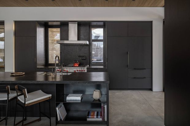 Dark, sophisticated contemporary kitchen with full-height charcoal cabinetry, a large island with integrated open shelving and seating, black benchtops, a cooking range with dark splashback, and tall windows. The space has a warm timber-lined ceiling and large pale stone-look floor tiles, creating a high-contrast, refined interior.