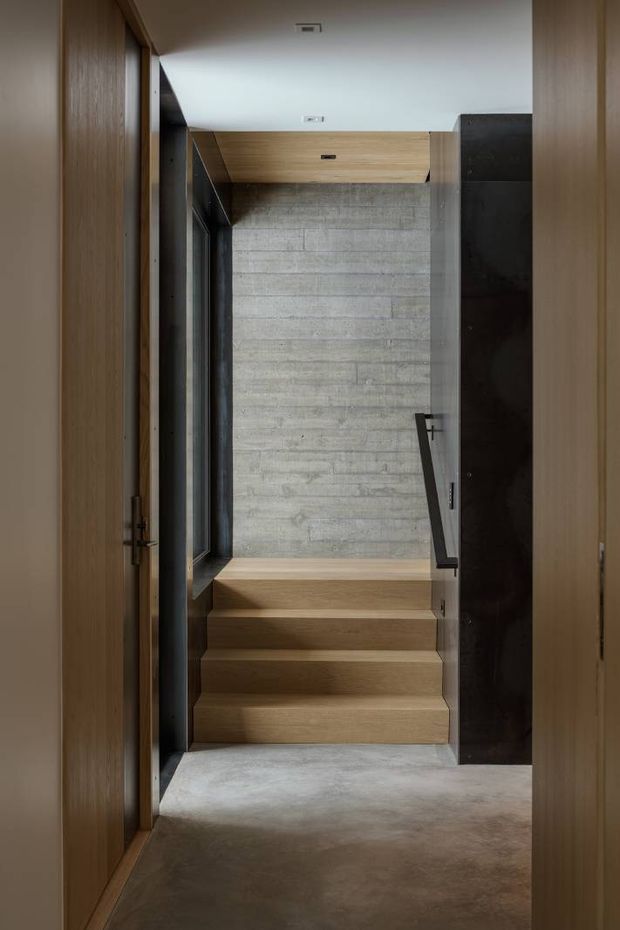 A narrow, dimly lit interior stairway viewed through timber-framed openings. The stair treads and surrounding ceiling are clad in warm timber, while the stairwell terminates at a textured grey concrete-block feature wall. Dark metal framing and a simple black handrail provide contrast, creating a restrained contemporary architectural composition.