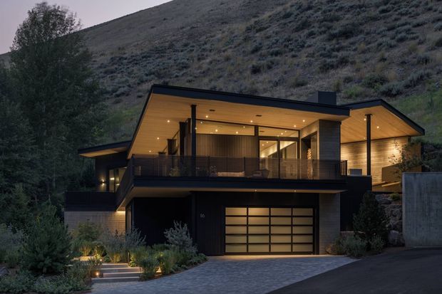 A contemporary multi-level hillside residence photographed at dusk. The home features a dark charcoal exterior, warm timber soffits and wall cladding, expansive glazed openings, a broad upper-level terrace with glass or slim dark balustrades, and an integrated double garage. Landscape planting and a stepped entry sit in front of the house, with a steep vegetated slope rising behind it.