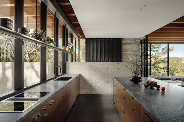 A spacious contemporary kitchen with a long island and separate perimeter cabinetry, dark stone benchtops, warm timber drawer fronts, a dark floor, and a concrete-look backsplash and partial-height wall. Full-height black-framed glazing overlooks trees and hills, while timber-lined ceiling sections and black vertical feature cladding add architectural detail. The kitchen has a calm, refined indoor-outdoor connection and abundant natural light.