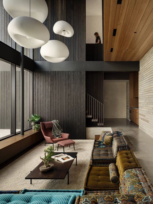 A dramatic double-height living room with a dark vertical timber feature wall, exposed timber ceiling, pale brick wall, black structural beams, and floor-to-ceiling glazing. The space is furnished with a large patterned sectional sofa, mid-century-style lounge chair and ottoman, low dark coffee tables, and oversized sculptural white pendant lights. A stairway and upper-level opening are visible at the rear, creating a strong sense of vertical volume.