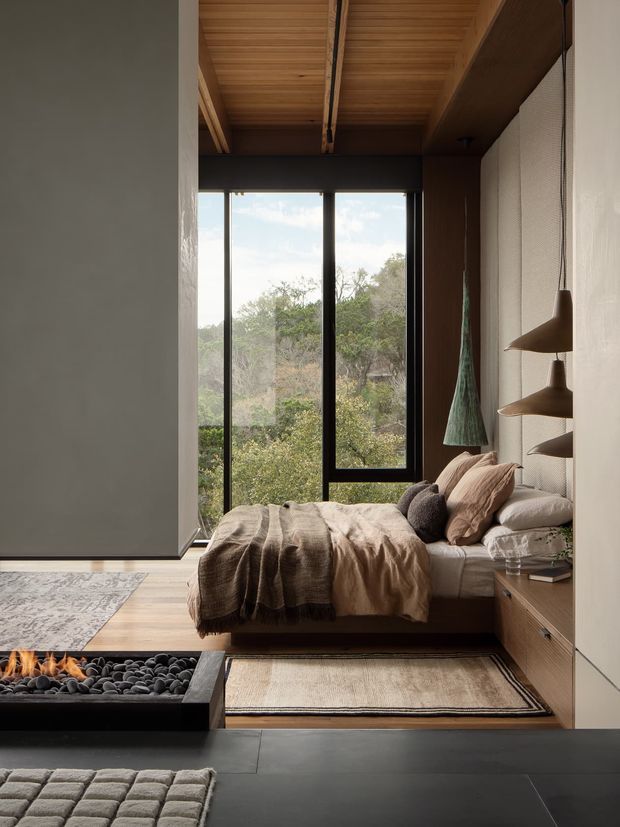 A refined, compact bedroom with a low platform bed positioned beside full-height black-framed glazing overlooking dense greenery. The room features a warm timber slatted ceiling, dark timber wall and built-in bedside joinery, a textured light wall panel, layered neutral bedding, sculptural hanging pendant lights, rugs, and a built-in linear fireplace in the foreground. The palette is earthy and muted, with a strong connection to the surrounding landscape.