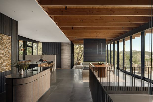 A sophisticated open-plan kitchen and living space with a long island, dark built-in cabinetry, timber bar seating, and a dining area beside extensive floor-to-ceiling glazing. A warm timber-lined ceiling with exposed beams spans the room, contrasting with dark vertical wall cladding, polished concrete flooring, and black metal balustrades. The glazing overlooks a leafy landscape, while a stair opening and railing define the circulation zone.