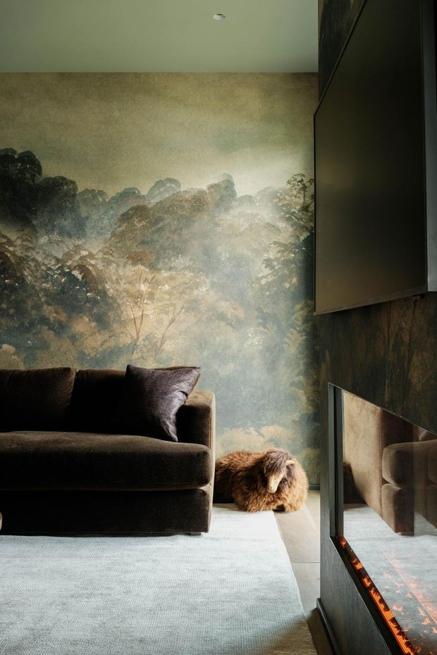A moody living room corner with a dark upholstered sofa, a large scenic forest mural or wallpapered feature wall, and a low built-in fireplace beneath a wall-mounted television. A small dog rests beside the sofa. The space uses dark timber-toned finishes, soft neutral carpet, and warm firelight, creating an intimate, cinematic atmosphere.
