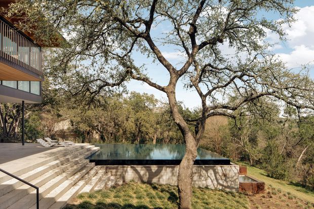 Exterior architectural view of a contemporary, multi-level home integrated into a wooded landscape. The scene features a cantilevered or elevated upper-level facade with extensive glazing and timber soffit, broad pale stone or concrete steps leading to a dark-edged rectangular reflecting or swimming pool, and a textured masonry retaining wall. Mature trees dominate the foreground and surround the building, creating a private, natural setting.