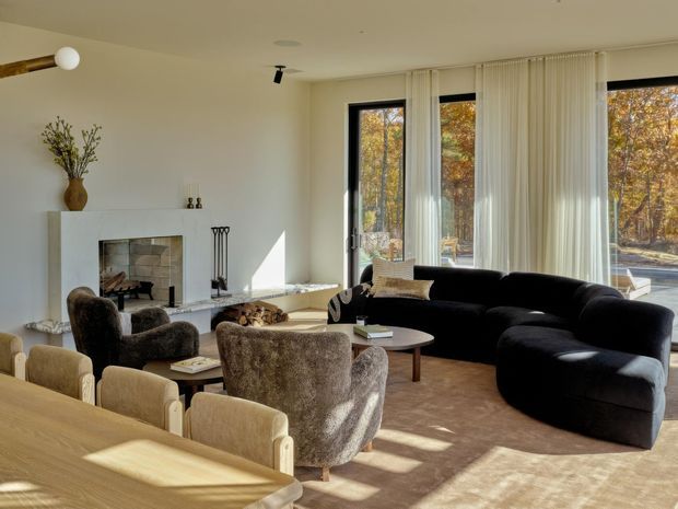 Sunlit open-plan living and dining area with a warm neutral palette. The room features a large curved dark sofa, textured lounge chairs, a timber dining table with upholstered chairs, a white fireplace surround with stacked-stone hearth, expansive black-framed glazing, sheer curtains, and a sculptural wall-mounted light. Autumn trees are visible outside.