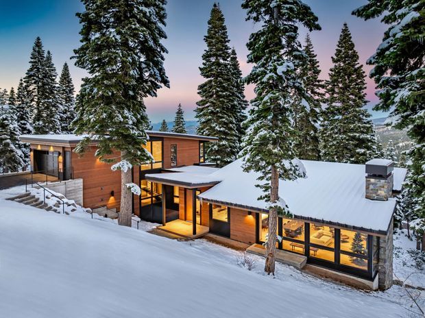Exterior view of a contemporary mountain lodge nestled among snow-covered evergreen trees on a steep snowy site. The low, interconnected building has warm timber cladding, expansive dark-framed glazing, broad snow-covered pitched roofs, and a stone chimney. Warm interior lighting is visible through the windows, creating a welcoming alpine atmosphere.