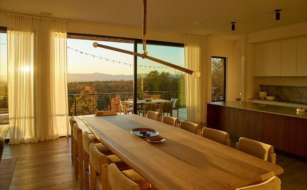 Warm, spacious open-plan dining and kitchen interior with a long timber dining table, upholstered dining chairs, and a contemporary kitchen with dark wood cabinetry and a pale stone benchtop. Full-height glazing and sheer curtains open onto a balcony or terrace with a distant landscape view. A linear timber-and-brass pendant light spans the dining table, while integrated ceiling spotlights provide additional illumination.