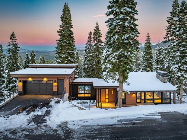 Snow-covered single-storey mountain home surrounded by tall evergreen trees, viewed from the driveway. The residence has a low-pitched roof, dark timber-toned cladding, extensive glazing, a projecting garage, and warm interior lighting at dusk. A snowy mountainous landscape is visible beyond the property.