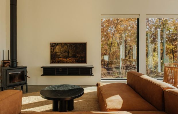 Warm contemporary living room with a freestanding black wood-burning stove and flue, a wall-mounted dark media console, framed artwork, a round dark coffee table, and a brown sectional sofa. Tall windows provide views of autumn trees and strong natural light, creating a cozy, refined atmosphere.