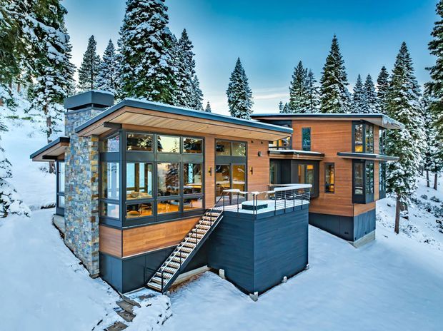 Exterior view of a modern multi-level mountain house surrounded by snow-covered evergreen forest. The home combines warm timber cladding, extensive dark-framed glazing, a low-pitched roof, and a prominent irregular natural-stone wall. Exterior stairs lead to an elevated entrance and terrace, with snow covering the surrounding site.