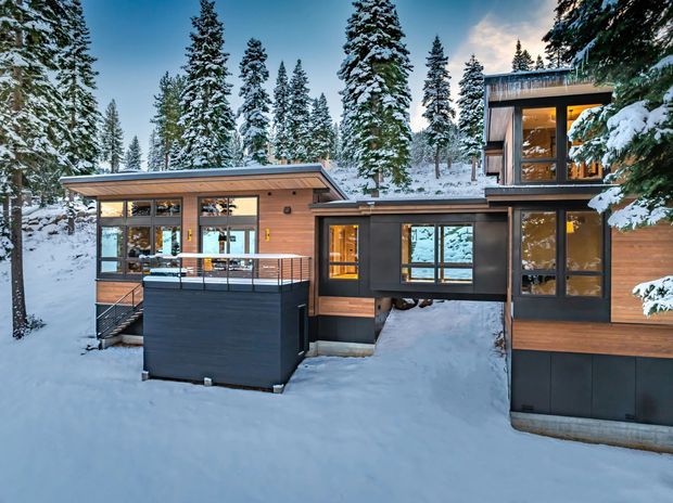 Exterior view of a contemporary multi-level mountain home set among snow-covered evergreen trees. The building has a low-pitched roof, extensive floor-to-ceiling glazing, warm timber-clad walls, dark metal or painted lower wall panels, and a raised deck or balcony with metal balustrades. Warm interior lighting is visible through the windows, while deep snow surrounds the structure.