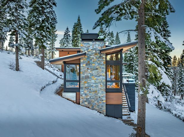 A contemporary mountain cabin set on a steep, snow-covered forested site. The compact multi-level home has angular shed-style roof forms, extensive glazing, warm timber cladding, dark metal trim and a prominent stacked-stone chimney or feature wall. Snow-covered conifers surround the building.