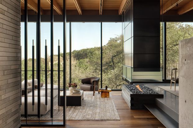 A sophisticated open-plan living room with floor-to-ceiling black-framed glazing overlooking a wooded landscape. The room features a large suspended contemporary fireplace with a stone hearth, dark built-in cabinetry, timber flooring, a warm timber ceiling with exposed beams, a neutral sectional sofa, armchairs, and a textured area rug. A tiled or masonry feature wall and concrete elements add architectural contrast.