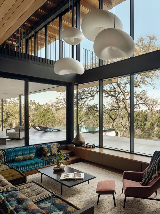 A spacious, double-height living room with floor-to-ceiling glazing opening onto a pool terrace and wooded landscape. The room features dark structural steel framing, a warm timber ceiling, a mezzanine-like upper-level railing, sculptural cream pendant lights, built-in timber window seating/storage, and richly patterned mid-century-style sofas and armchairs arranged around a low black coffee table.