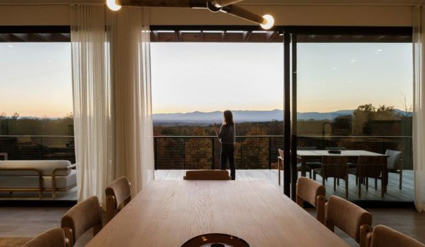 A warm, contemporary open-plan dining and living space with a large timber dining table in the foreground, an upholstered sofa to the left, and full-height glazed sliding doors opening to a balcony or terrace. The room has exposed timber ceiling elements, slim dark window frames, sheer curtains, and pendant-style linear lighting. A person stands outside on the terrace, with expansive distant hills visible beyond the railing.