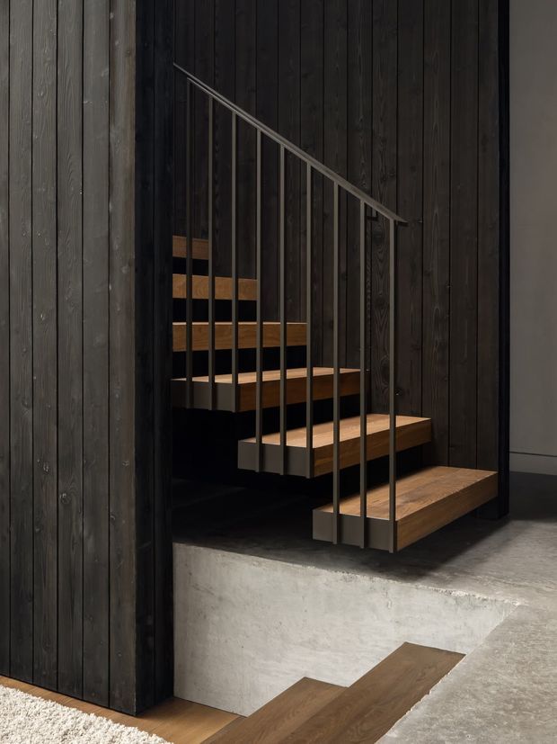 A dramatic minimalist stairway with floating timber treads supported within a dark vertical timber-lined enclosure. Slim black metal balusters and handrail run diagonally along the stairs. The stair opening sits above a raw concrete plinth, with polished concrete flooring and dark timber wall cladding creating a high-contrast, moody architectural interior.