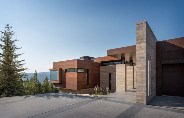 A contemporary multi-level residence viewed from the driveway, composed of strong rectilinear volumes with extensive timber-toned cladding, concrete walls, deep roof overhangs, and dark-framed glazing. The home sits in a wooded, mountainous setting, with a broad paved approach and an integrated garage at right. Clear blue sky and evergreen trees frame the angular facade.