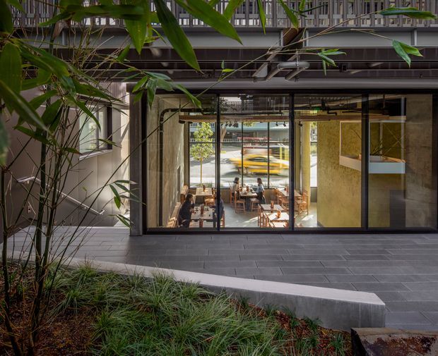 A contemporary commercial-style building courtyard viewed from a landscaped outdoor area. Large black-framed glazed doors and windows open into a bright dining or lounge space, with exposed structural elements and an upper-level balcony or walkway. Dark grey tiled paving, textured concrete walls, integrated bench-like edging, and dense bamboo-style planting create a modern indoor-outdoor setting.