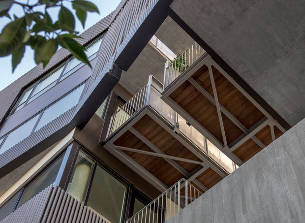 Upward-looking architectural view of a contemporary multi-storey building with projecting balconies, exposed structural framing, timber soffits, metal balustrades, corrugated vertical facade elements, extensive glazing, and a raw concrete side wall. Vegetation partially frames the upper left edge.
