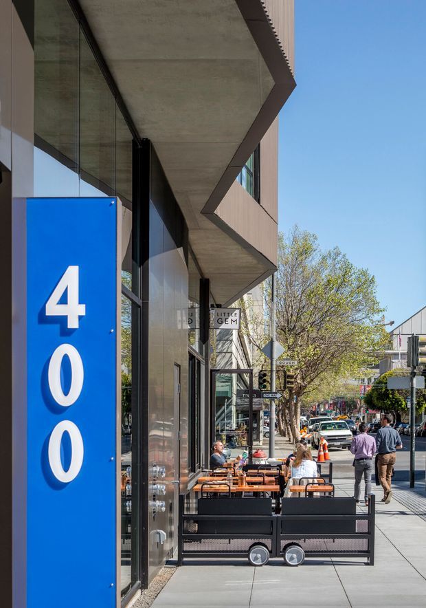 Street-level view of a contemporary commercial building facade with a prominent blue vertical address sign reading 400, full-height glazing, angular projecting concrete canopy elements, and ground-floor cafe seating extending along the wide sidewalk. The scene is urban, bright, and people-oriented, with neighbouring storefronts and street trees visible beyond.