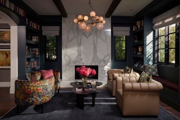 A richly styled, moody living room with dark navy built-in shelving, a dramatic veined marble fireplace surround, exposed dark ceiling beams, and large black-framed windows. The room contains a sculptural globe chandelier, a tan tufted leather sofa, a colourful floral curved lounge chair, a dark coffee table, patterned cushions, and a deep charcoal rug.