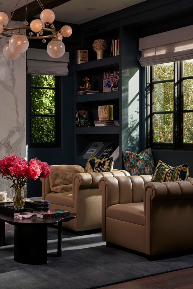 A richly layered, moody living room with deep charcoal-blue walls, built-in open shelving, black-framed windows, and soft grey Roman blinds. Two rounded tan upholstered armchairs and a matching sofa surround a low black coffee table on a dark rug. Patterned cushions, books, decorative objects, a large marble-look wall panel, and a sculptural globe chandelier add visual interest, while daylight filters through the windows and greenery is visible outside.