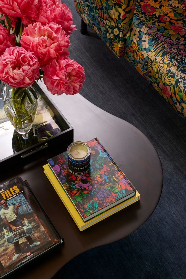 Close-up styled living room vignette featuring a dark oval coffee table with stacked colourful art books and a candle, a mirrored tray holding a glass vase of bright pink peonies, and a richly patterned floral upholstered sofa. A dark textured floor and dramatic, saturated colour palette create a bold, luxurious atmosphere.