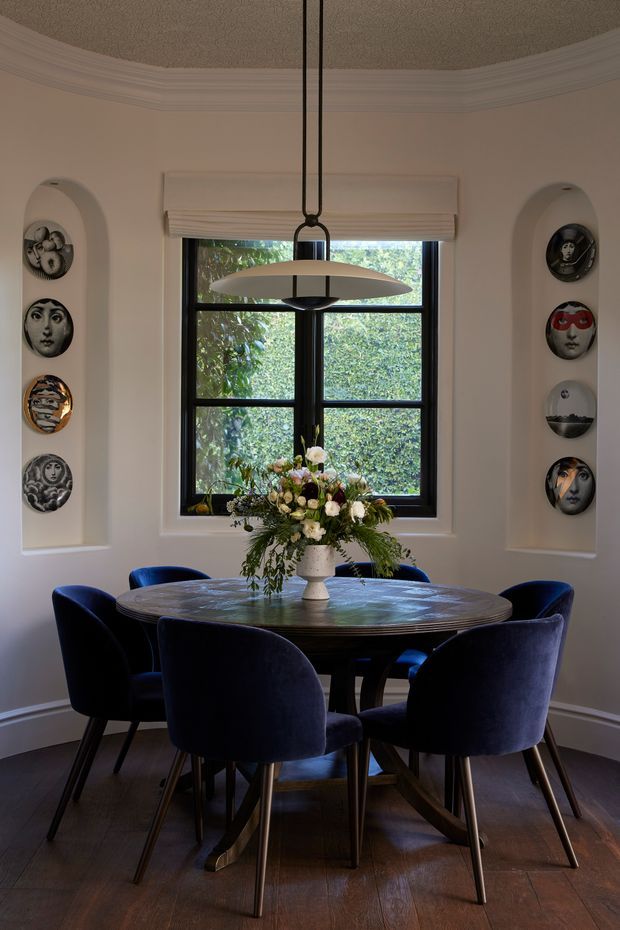 A refined dining room with a round dark wood dining table surrounded by deep navy upholstered chairs. A sculptural pendant light hangs above a floral centrepiece, while a black-framed window provides a leafy garden view. Curved wall niches display decorative plates, and ornate ceiling cornices and dark timber flooring add a traditional architectural character.