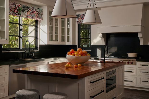 A bright, traditional-style kitchen with white Shaker cabinetry, black-framed windows, a dark backsplash, and a large central island topped with warm timber. The island includes seating, drawers, an integrated oven, and a sink with a gooseneck tap. Patterned Roman blinds, pendant lights, and a substantial painted fireplace surround add character, while sunlight and greenery are visible through the windows.