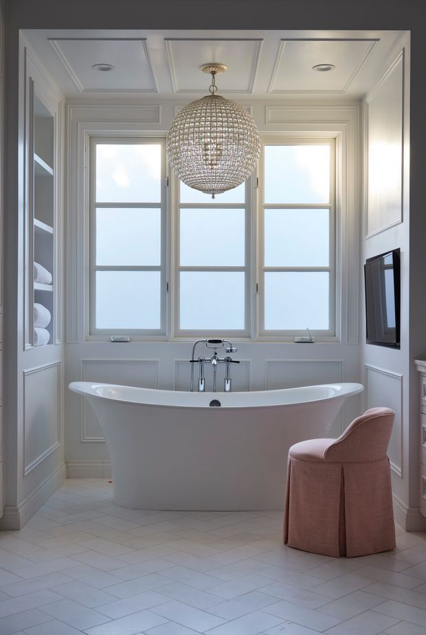 A bright, elegant bathroom centered on a freestanding white bathtub beneath three tall frosted-glass windows. The room features white decorative wall panelling, a coffered ceiling with recessed lights, a large spherical pendant light, pale tiled flooring, built-in shelving with folded towels, and a wall-mounted television. A blush-pink upholstered chair adds a soft accent.
