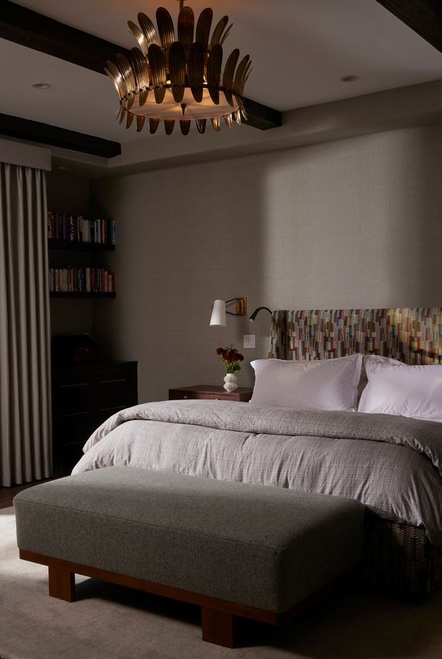 A moody, refined bedroom with a large upholstered bed dressed in pale grey bedding, a colourful patterned headboard, and a matching upholstered bench with a timber base. Dark built-in shelving and cabinetry sit beside the bed, with curtains, textured wallcovering, dark exposed ceiling beams, and a dramatic sculptural brass-and-dark chandelier. Warm accent lighting and layered textiles create an intimate, luxurious atmosphere.