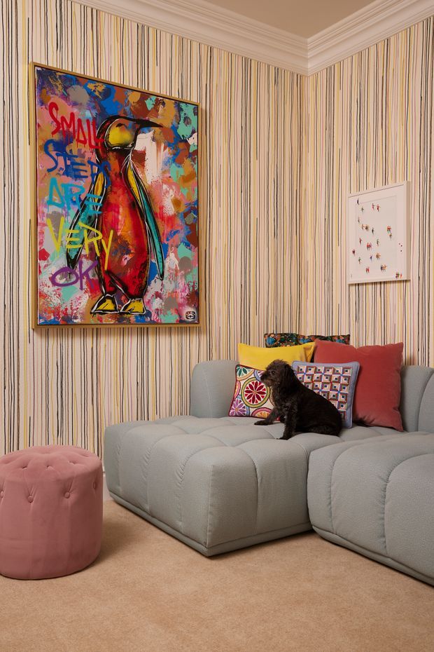 A richly styled living room corner featuring a large pale grey sectional sofa, colourful patterned cushions, and a pink tufted ottoman on a beige carpet. The walls are covered in bold vertically striped wallpaper with ornate white crown moulding. Colourful contemporary artwork and a smaller framed artwork add playful visual accents, while a small dark dog sits on the sofa.