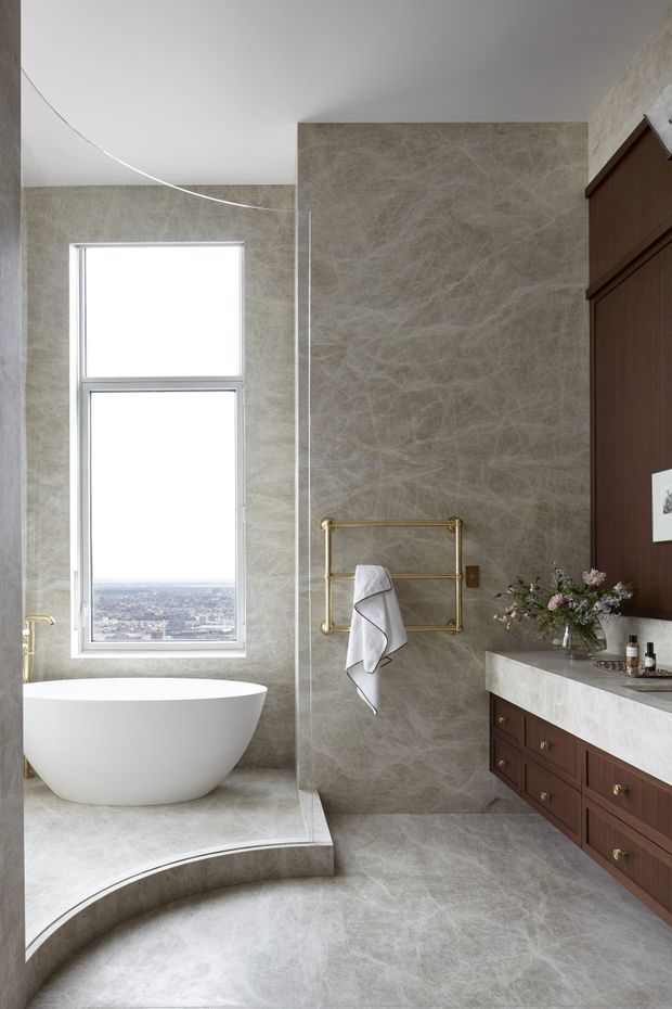 A refined contemporary bathroom with a freestanding white soaking tub positioned beside a large window, enclosed by a curved glass shower screen. The space is finished in continuous pale grey-beige veined stone across the walls and floor, with dark walnut-toned vanity cabinetry, a pale stone countertop, and brass towel rails and fittings. A city view is visible through the window.
