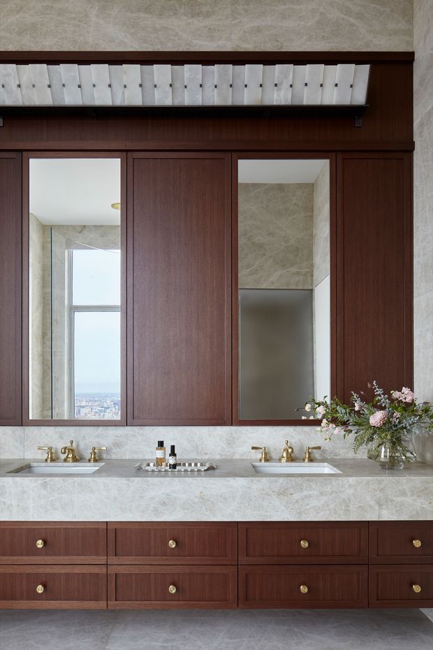 A refined double-sink bathroom vanity with extensive dark timber cabinetry, pale veined stone benchtop and backsplash, brass fixtures, and framed mirrors. A narrow upper shelf displays evenly arranged decorative objects, while a floral arrangement adds softness. A city view is visible through the reflected window.